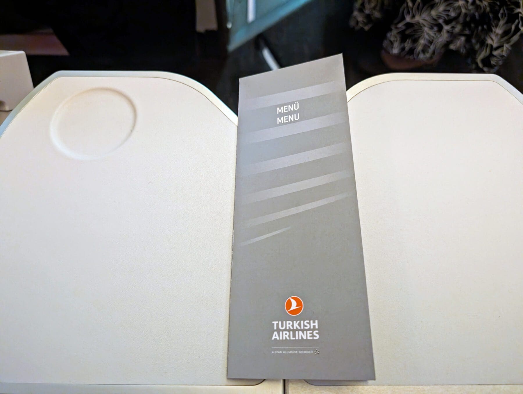 Turkish Airlines Food Review Economy Class 9 A Turkish Airlines menu rests on a tray table, featuring a gray cover with the airline's logo in orange and white text. Inside, passengers can explore the exquisite selection of Turkish Airlines meals, promising a delightful culinary journey at 30,000 feet.