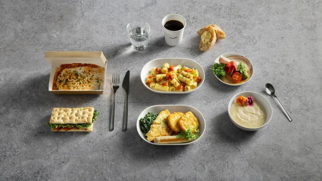 Finnair's Winter Menu Leans on Seasonal Finnish Ingredients 3 A meal set on a grey surface featuring pasta, bread, salad, sandwich, egg dish, yogurt with fruitโsimilar to the variety found in Air Astana kids packsโplus a cup of coffee and a glass of water.
