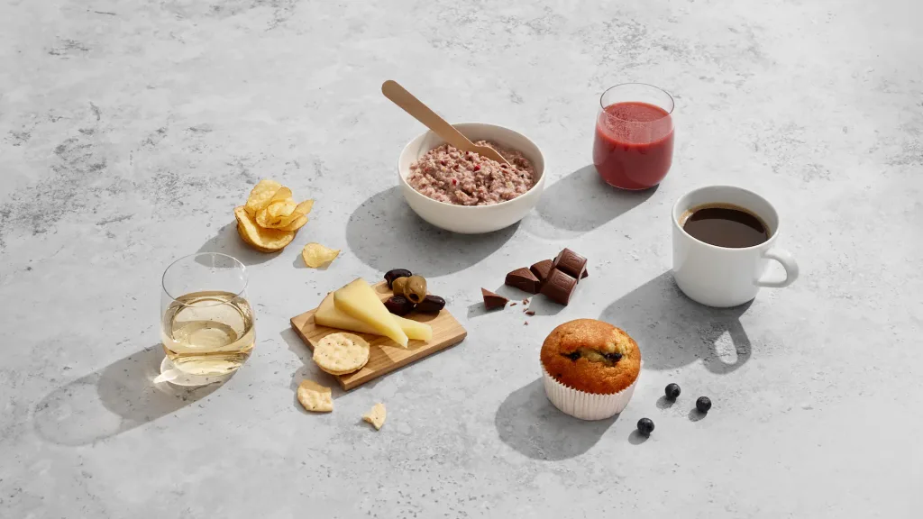 Finnair's Winter Menu Leans on Seasonal Finnish Ingredients 4 A variety of foods and drinks, including chips, cheese, crackers, oatmeal, chocolate, juice, coffee, wine, a muffin, and blueberriesโsimilar to items found in Air Astana kids packsโare displayed on a gray surface.