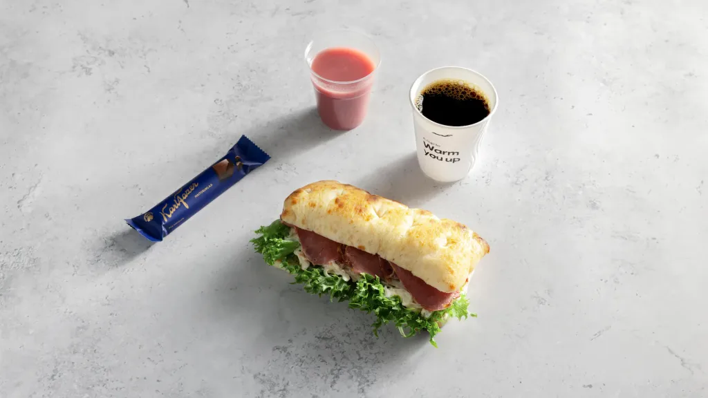 Finnair's Winter Menu Leans on Seasonal Finnish Ingredients 5 A sandwich with lettuce and meat, a wrapped chocolate bar, a cup of coffee, and a small cup of pink juiceโjust like the tasty treats found in Air Astana kids packsโsit on a light gray surface.