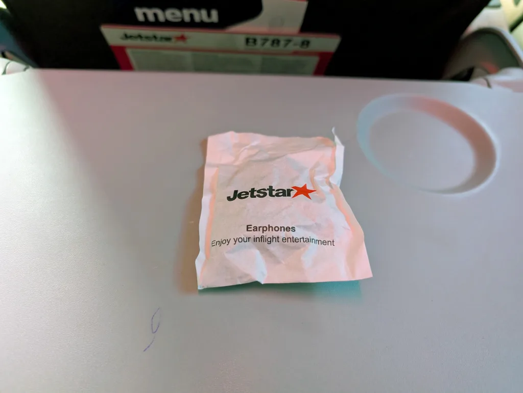 Jetstar Economy Review: Melbourne to Singapore 13 A crumpled Jetstar earphones packet sits on an airplane tray table, with a seat back and menu visible in the background.