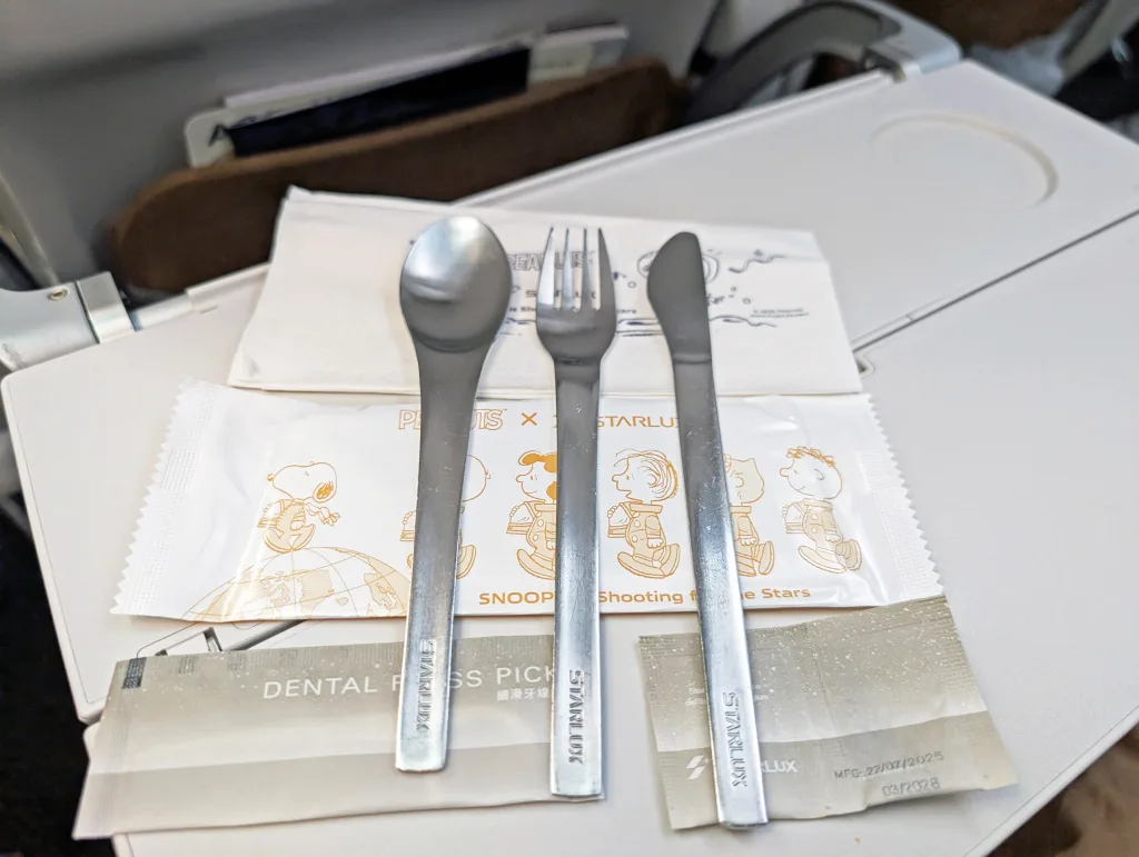Starlux Airlines Review 2025: Taipei to Singapore 8 A set of metal spoon, fork, and knife is arranged on an airplane tray table, along with napkins, a dental floss pick, and a Snoopy-themed packaged item.