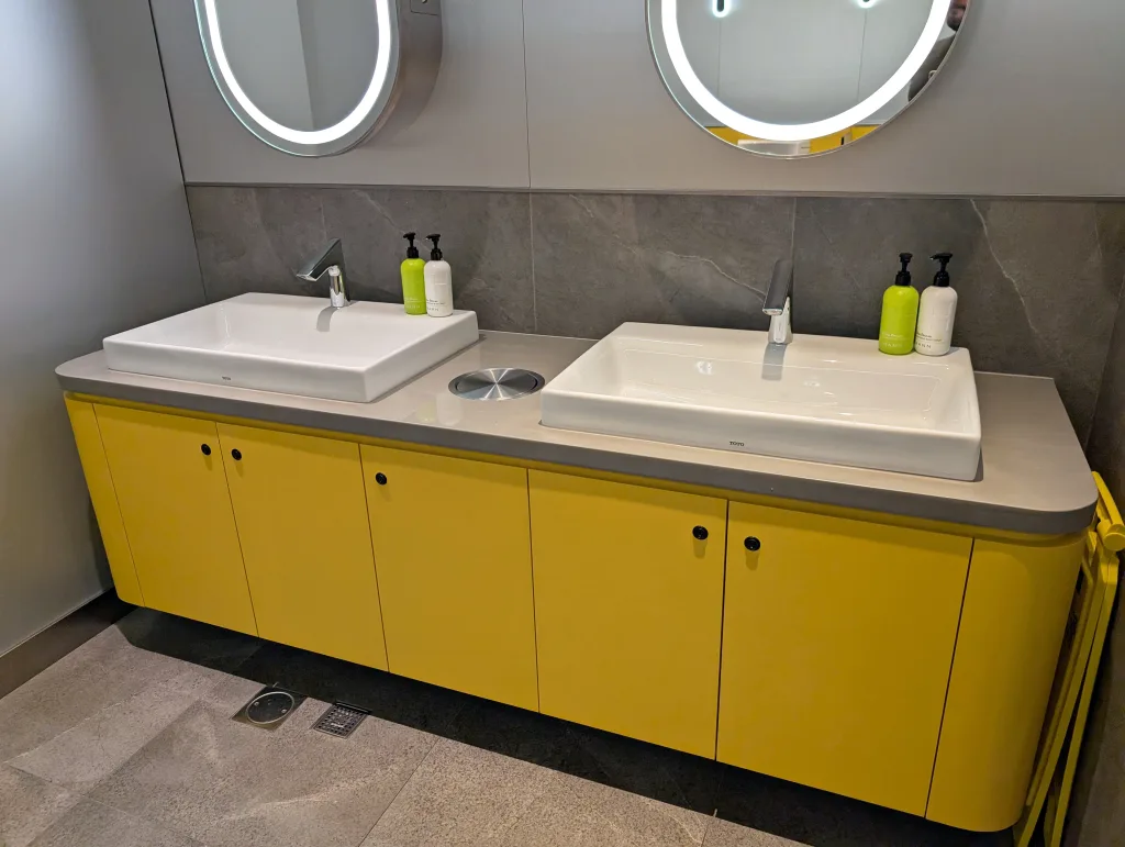 Starlux Airlines Review 2025: Taipei to Singapore 26 Two rectangular sinks with faucets are set in a yellow vanity with gray countertop, under two round mirrors in a modern bathroom. Soap dispensers are placed beside each sink.