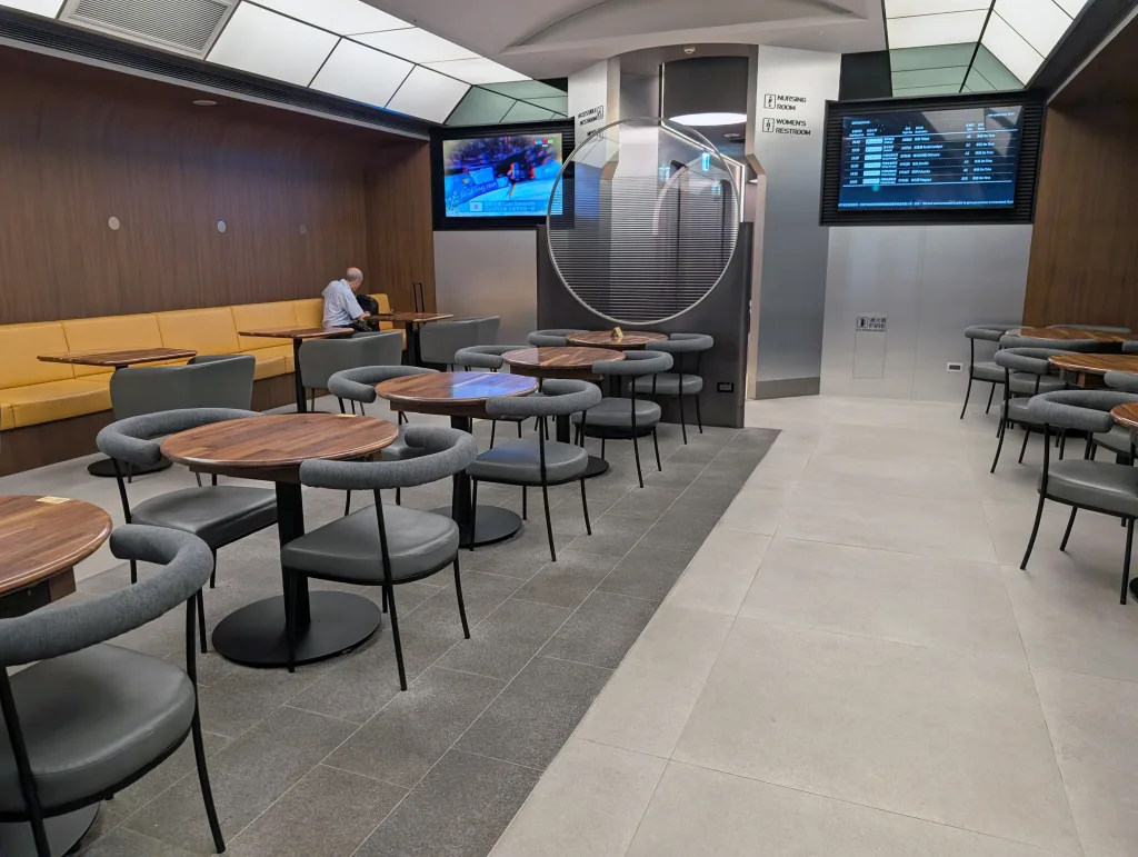 Starlux Airlines Review 2025: Taipei to Singapore 17 Modern airport lounge with empty tables and chairs, one person seated at the back, wall-mounted screens displaying sports and flight information.