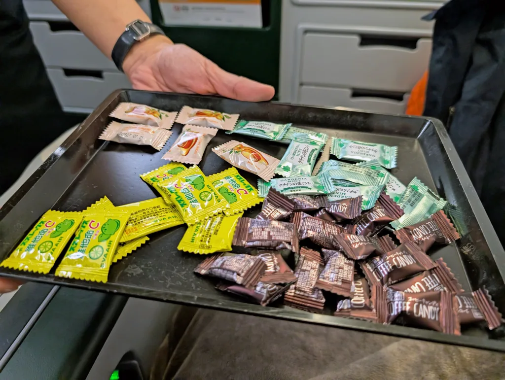 A person holds a tray containing various individually wrapped sweets in yellow, brown, green, and beige wrappers.