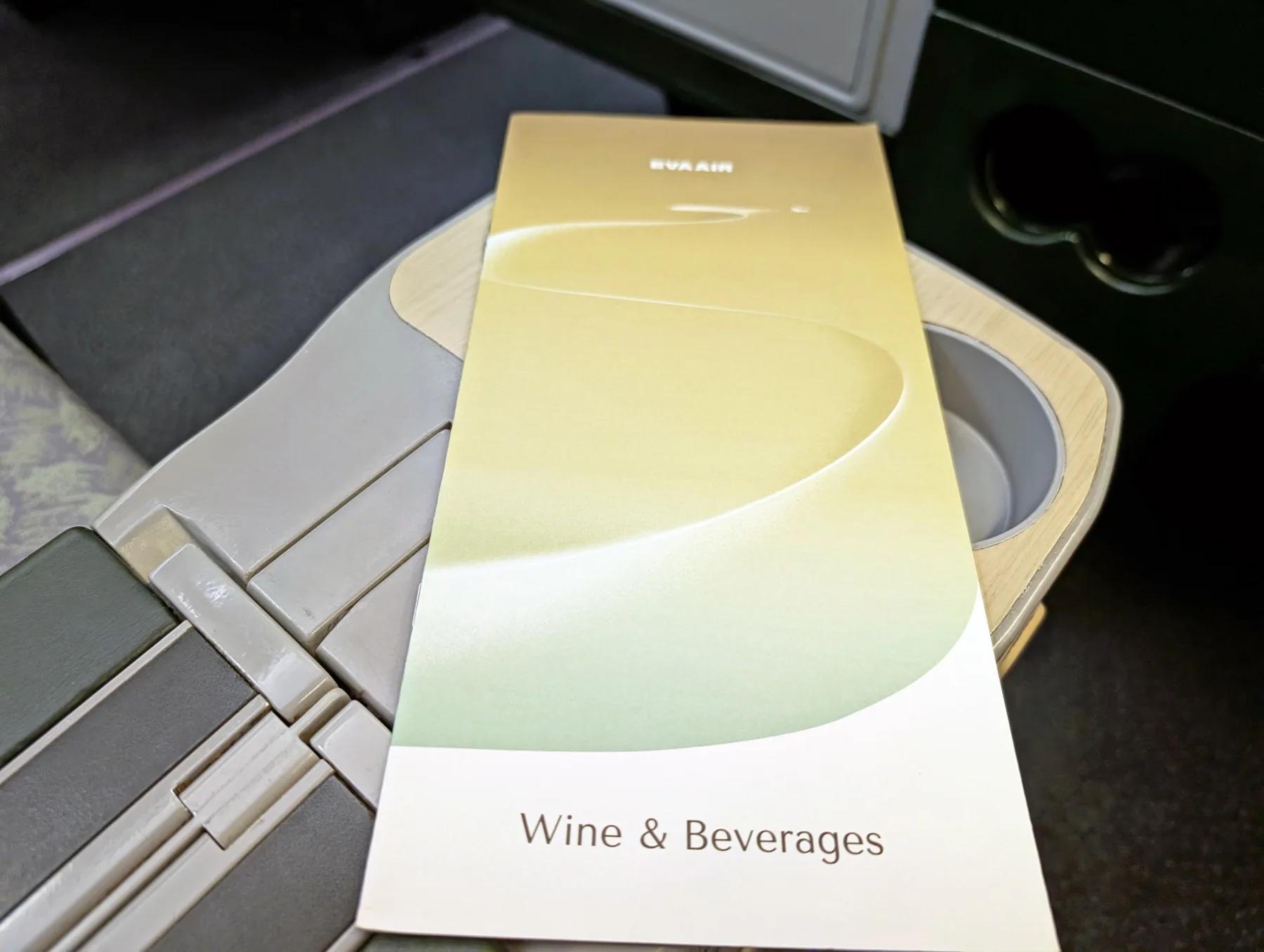 A wine and beverages menu with “EVA AIR” on the cover is placed on an aeroplane tray table near a cup holder.