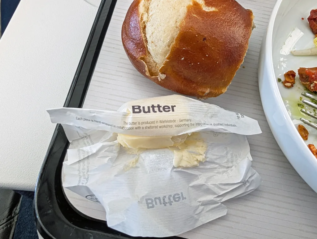 Lufthansa: Paris to Toulouse 5 An opened butter packet with some butter remaining sits next to a bread roll on a tray, with part of a plate of food and cutlery visible.
