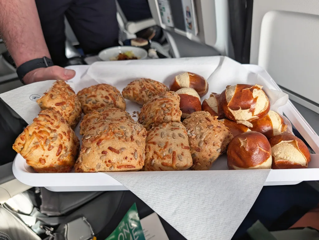 Lufthansa: Paris to Toulouse 4 A tray with cheese-topped bread rolls and small pretzel rolls is being served on an aeroplane, with serviettes under the bread.