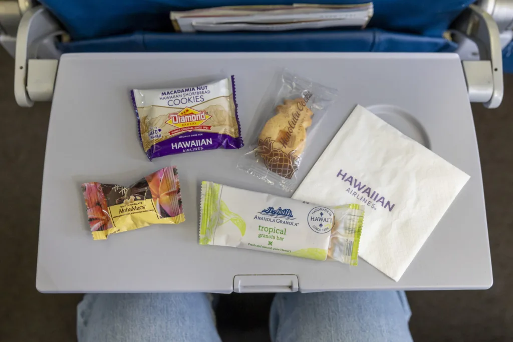 Chef Sheldon Simeon brings Tin Roof to the skies on Hawaiian Airlines 12 An aeroplane tray table with packaged snacks crafted by Hawaiian Airlines chef Sheldon Simeon, a serviette labelled "Hawaiian Airlines," and a passenger's legs partially visible below.