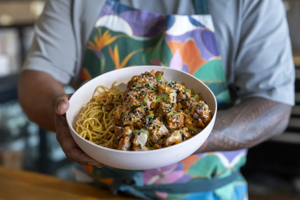 Chef Sheldon Simeon brings Tin Roof to the skies on Hawaiian Airlines 11 A person in a colourful apron holds a bowl of noodles topped with seasoned chicken pieces and garnished with herbs and sesame seeds.