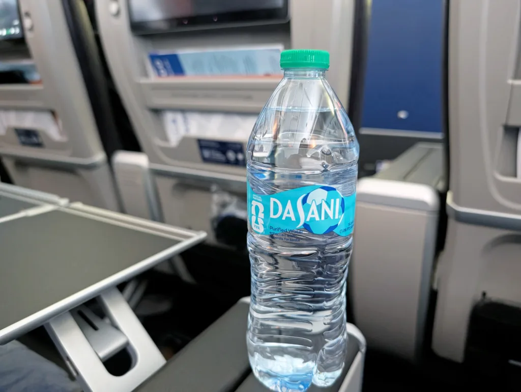 A bottle of Dasani water sits on a tray table in an aeroplane cabin, with seatback screens and pockets visible in the background.